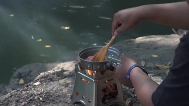 Bushcraft konsepti basit bir odun sobası kullanarak nehir kenarında sandviç ve sıcak çay hazırlıyor. Erkek bir maceracı, yemyeşil ağaçlarla çevrili temiz bir nehrin kenarında kum cadıları ve kaynar su pişiriyor.