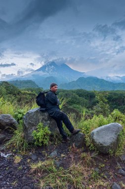 Bir maceracı öğleden sonra bir kayanın üzerinde oturmuş bulutlu bir dağa bakıyordu. Asyalı adam, arka planda Merapi Dağı olan bir kayanın üzerinde oturarak doğanın huzurunun tadını çıkarıyor.