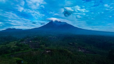 Merapi Dağı 'nın havadan çekilmiş fotoğrafı. Karanlık bir sabahta etrafındaki bulutlar açıkça görülüyor. Önünde sık bir orman bulunan güneyden aktif volkan manzarası
