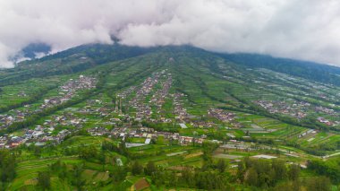 Aerial photo from the foot of the mountain with rice fields and several villages quite close to the top of the mountain. Aerial panorama of the village area and rice fields at the foot of Mount Merapi clipart