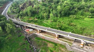 Her iki tarafında vadiler ve tepeler olan yeşil pirinç tarlalarını ayıran bir köprünün hava görüntüleri. Endonezya kırsalındaki tepeleri ve pirinç tarlalarını kesen otobanın hava manzarası