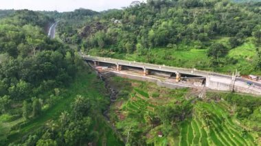 Her iki tarafında vadiler ve tepeler olan yeşil pirinç tarlalarını ayıran bir köprünün hava görüntüleri. Endonezya kırsalındaki tepeleri ve pirinç tarlalarını kesen otobanın hava manzarası