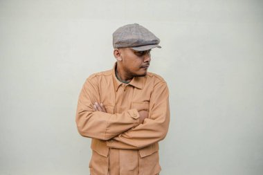 Bronzlaşmış tenli yetişkin bir adam krem renkli bir arka plana karşı poz verir. Yetişkin bir Güneydoğu Asyalı 'nın yarı boy portresi. Gazeteci şapkası ve kahverengi ceket giyiyor.