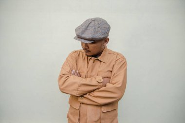 Bronzlaşmış tenli yetişkin bir adam krem renkli bir arka plana karşı poz verir. Yetişkin bir Güneydoğu Asyalı 'nın yarı boy portresi. Gazeteci şapkası ve kahverengi ceket giyiyor.