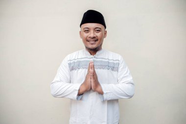 Müslüman bir adamın Kurban Bayramı 'nda özür dileme jesti yaparken çekilmiş ayrı bir konsept fotoğrafı. Beyaz Müslüman gömlekli ve siyah şapkalı Asyalı bir adam beyaz arka planda af diliyor..