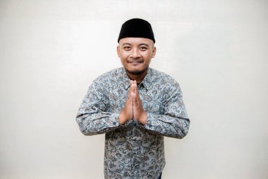 Müslüman bir adamın şal desenli gömlek giydiği ve siyah şapkalı bir adamın bayramda özür jesti yaptığı ayrı bir konsept fotoğrafı. Batik gömlekli ve şapkalı Asyalı bir adam beyaz bir arka plana karşı merhamet dileniyor.