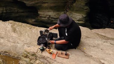 Erkek bir maceracı nehir kıyısındaki antik kayalarda baharatlı çay yapmak için su kaynatır. Bushcraft konsepti, volkanik lavlardan antik kayalarda sıcak çay yaparak doğanın huzurunun tadını çıkarmaktır..
