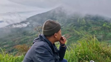 Asyalı bir yolcunun açık hava aktiviteleri V60 kahve içip tepenin zirvesinde dumanlı bir dağ arkası ile sigara içmesi. Erkek bir maceraperest sabahları zirvede sıcak bir içkinin tadını çıkarıyor.