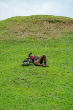 Bir maceracı yeşil çimenlerin üzerinde uzanarak dinleniyor. Asyalı adam, güzel ve temiz yeşil bir tepede dinlenip uzanarak doğanın huzurunun tadını çıkarıyor..