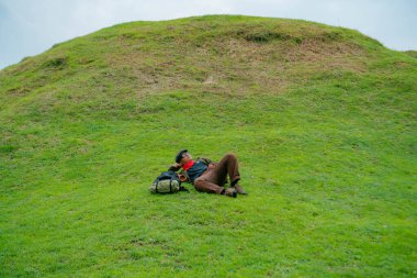 Bir maceracı yeşil çimenlerin üzerinde uzanarak dinleniyor. Asyalı adam, güzel ve temiz yeşil bir tepede dinlenip uzanarak doğanın huzurunun tadını çıkarıyor..