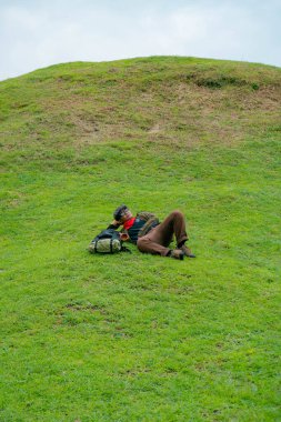 Bir maceracı yeşil çimenlerin üzerinde uzanarak dinleniyor. Asyalı adam, güzel ve temiz yeşil bir tepede dinlenip uzanarak doğanın huzurunun tadını çıkarıyor..