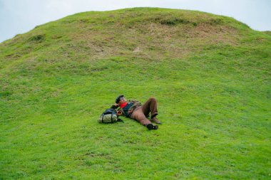 Bir maceracı yeşil çimenlerin üzerinde uzanarak dinleniyor. Asyalı adam, güzel ve temiz yeşil bir tepede dinlenip uzanarak doğanın huzurunun tadını çıkarıyor..