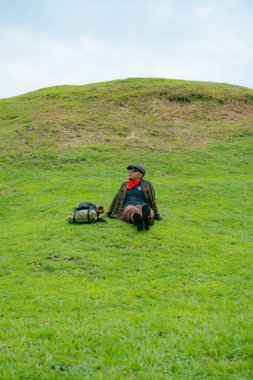 Bir maceracı yeşil çimlerin üzerinde oturarak dinleniyor. Asyalı adam gömülü bir Budist tapınağı olduğu anlaşılan yeşil bir tepede dinlenerek doğanın huzurunun tadını çıkarıyor..