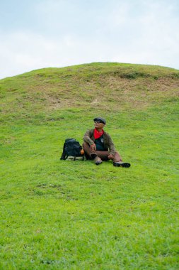 Bir maceracı yeşil çimlerin üzerinde oturarak dinleniyor. Asyalı adam gömülü bir Budist tapınağı olduğu anlaşılan yeşil bir tepede dinlenerek doğanın huzurunun tadını çıkarıyor..
