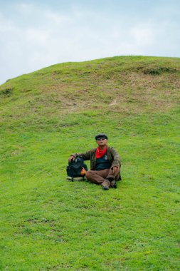 Bir maceracı yeşil çimlerin üzerinde oturarak dinleniyor. Asyalı adam gömülü bir Budist tapınağı olduğu anlaşılan yeşil bir tepede dinlenerek doğanın huzurunun tadını çıkarıyor..