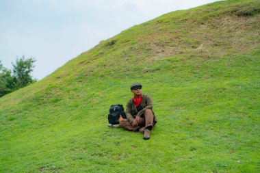 Bir maceracı yeşil çimlerin üzerinde oturarak dinleniyor. Asyalı adam gömülü bir Budist tapınağı olduğu anlaşılan yeşil bir tepede dinlenerek doğanın huzurunun tadını çıkarıyor..
