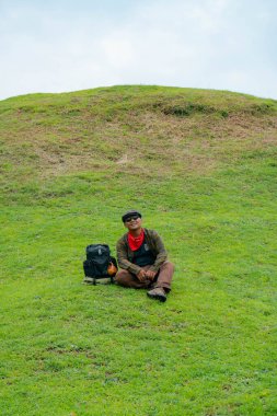 Bir maceracı yeşil çimlerin üzerinde oturarak dinleniyor. Asyalı adam gömülü bir Budist tapınağı olduğu anlaşılan yeşil bir tepede dinlenerek doğanın huzurunun tadını çıkarıyor..