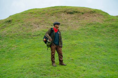 Erkek bir maceracı yeşil çimenlerin engin güzelliğinin tadını çıkarıyor. Asyalı erkek gezgin rahatlar ve temiz ve güzel yeşil tepelerin doğal cazibesine hayran kalır..