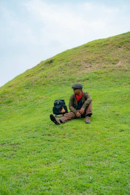 Bir maceracı yeşil çimlerin üzerinde oturarak dinleniyor. Asyalı adam gömülü bir Budist tapınağı olduğu anlaşılan yeşil bir tepede dinlenerek doğanın huzurunun tadını çıkarıyor..