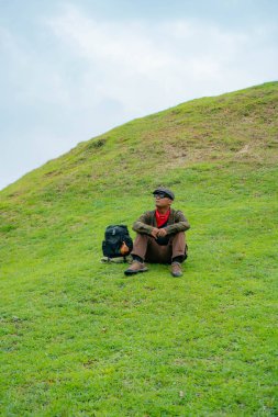 Bir maceracı yeşil çimlerin üzerinde oturarak dinleniyor. Asyalı adam gömülü bir Budist tapınağı olduğu anlaşılan yeşil bir tepede dinlenerek doğanın huzurunun tadını çıkarıyor..