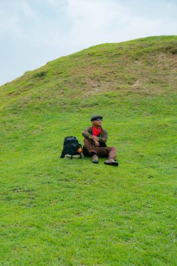 Bir maceracı yeşil çimlerin üzerinde oturarak dinleniyor. Asyalı adam gömülü bir Budist tapınağı olduğu anlaşılan yeşil bir tepede dinlenerek doğanın huzurunun tadını çıkarıyor..