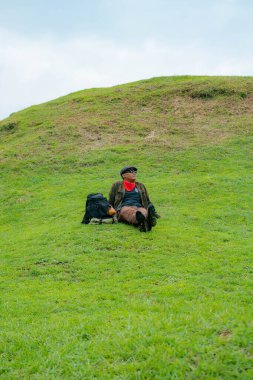 Bir maceracı yeşil çimlerin üzerinde oturarak dinleniyor. Asyalı adam gömülü bir Budist tapınağı olduğu anlaşılan yeşil bir tepede dinlenerek doğanın huzurunun tadını çıkarıyor..