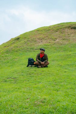 Bir maceracı yeşil çimlerin üzerinde oturarak dinleniyor. Asyalı adam gömülü bir Budist tapınağı olduğu anlaşılan yeşil bir tepede dinlenerek doğanın huzurunun tadını çıkarıyor..