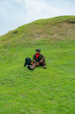 Bir maceracı yeşil çimlerin üzerinde oturarak dinleniyor. Asyalı adam gömülü bir Budist tapınağı olduğu anlaşılan yeşil bir tepede dinlenerek doğanın huzurunun tadını çıkarıyor..