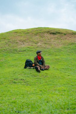 Bir maceracı yeşil çimlerin üzerinde oturarak dinleniyor. Asyalı adam gömülü bir Budist tapınağı olduğu anlaşılan yeşil bir tepede dinlenerek doğanın huzurunun tadını çıkarıyor..