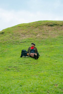 Bir maceracı yeşil çimlerin üzerinde oturarak dinleniyor. Asyalı adam gömülü bir Budist tapınağı olduğu anlaşılan yeşil bir tepede dinlenerek doğanın huzurunun tadını çıkarıyor..
