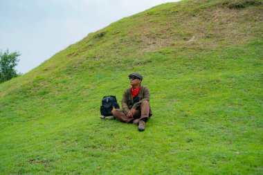 Bir maceracı yeşil çimlerin üzerinde oturarak dinleniyor. Asyalı adam gömülü bir Budist tapınağı olduğu anlaşılan yeşil bir tepede dinlenerek doğanın huzurunun tadını çıkarıyor..