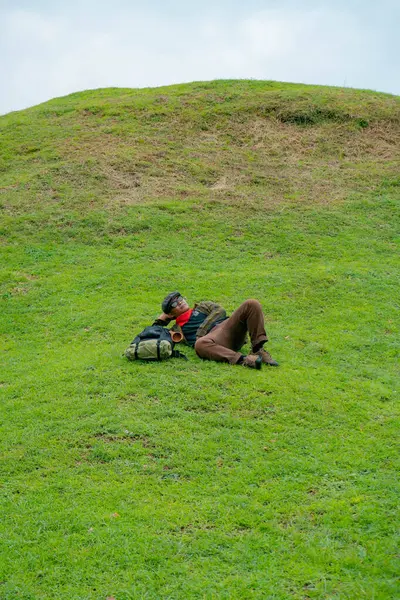 Bir maceracı yeşil çimenlerin üzerinde uzanarak dinleniyor. Asyalı adam, güzel ve temiz yeşil bir tepede dinlenip uzanarak doğanın huzurunun tadını çıkarıyor..