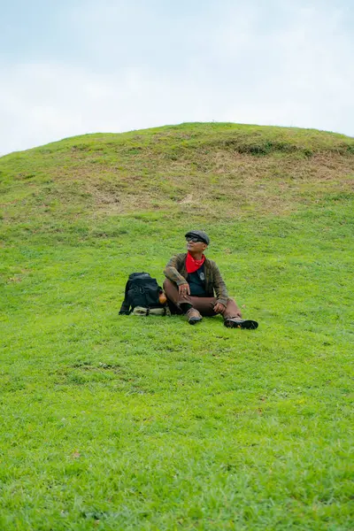 Bir maceracı yeşil çimlerin üzerinde oturarak dinleniyor. Asyalı adam gömülü bir Budist tapınağı olduğu anlaşılan yeşil bir tepede dinlenerek doğanın huzurunun tadını çıkarıyor..