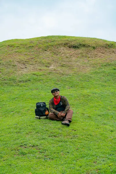 Bir maceracı yeşil çimlerin üzerinde oturarak dinleniyor. Asyalı adam gömülü bir Budist tapınağı olduğu anlaşılan yeşil bir tepede dinlenerek doğanın huzurunun tadını çıkarıyor..
