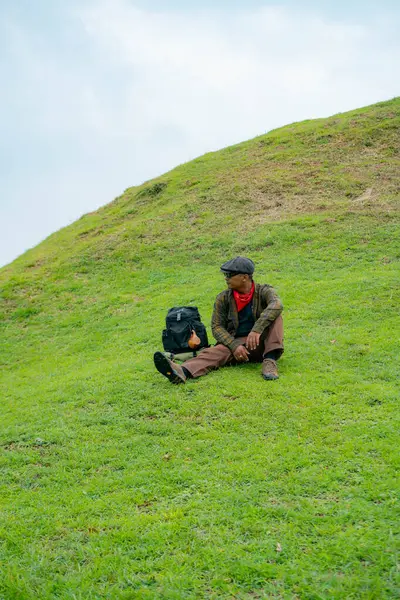 Bir maceracı yeşil çimlerin üzerinde oturarak dinleniyor. Asyalı adam gömülü bir Budist tapınağı olduğu anlaşılan yeşil bir tepede dinlenerek doğanın huzurunun tadını çıkarıyor..