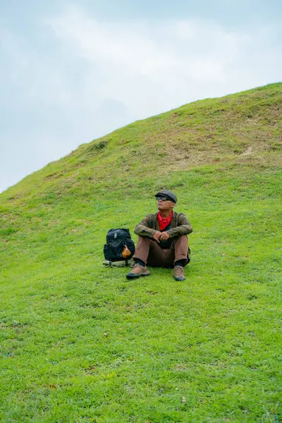 Bir maceracı yeşil çimlerin üzerinde oturarak dinleniyor. Asyalı adam gömülü bir Budist tapınağı olduğu anlaşılan yeşil bir tepede dinlenerek doğanın huzurunun tadını çıkarıyor..