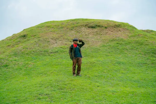 Erkek bir maceracı yeşil çimenlerin engin güzelliğinin tadını çıkarıyor. Asyalı erkek gezgin rahatlar ve temiz ve güzel yeşil tepelerin doğal cazibesine hayran kalır..