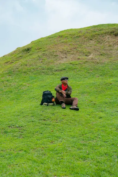 Bir maceracı yeşil çimlerin üzerinde oturarak dinleniyor. Asyalı adam gömülü bir Budist tapınağı olduğu anlaşılan yeşil bir tepede dinlenerek doğanın huzurunun tadını çıkarıyor..