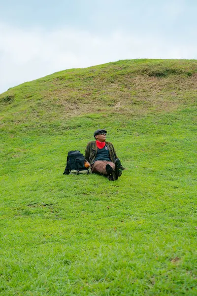 Bir maceracı yeşil çimlerin üzerinde oturarak dinleniyor. Asyalı adam gömülü bir Budist tapınağı olduğu anlaşılan yeşil bir tepede dinlenerek doğanın huzurunun tadını çıkarıyor..