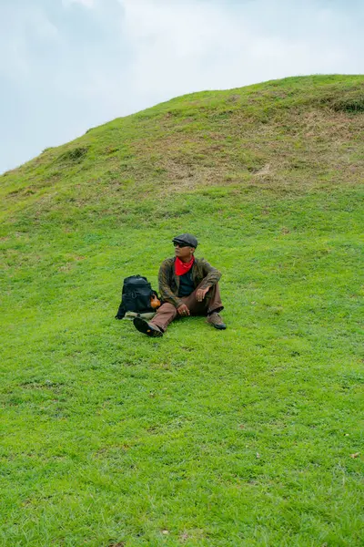 Bir maceracı yeşil çimlerin üzerinde oturarak dinleniyor. Asyalı adam gömülü bir Budist tapınağı olduğu anlaşılan yeşil bir tepede dinlenerek doğanın huzurunun tadını çıkarıyor..