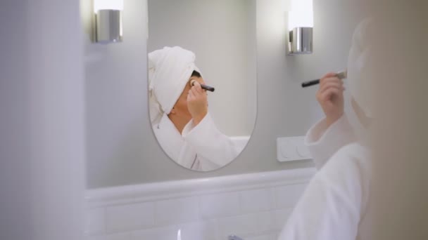 女の子が鏡の前に化粧をしています 若い女の子が化粧をする ホワイトバスルーム 女の子は顔に化粧をブラシで塗っています 女の子がシャワーを浴びてミーティングに行く — ストック動画
