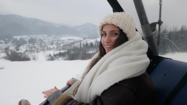 坐滑雪车的女孩 一个女孩倒在滑雪电梯上 滑雪电梯 年轻的女孩对着摄像机笑 寒假冬天 下雪了 一个穿冬衣的女孩 山区度假 — 图库视频影像