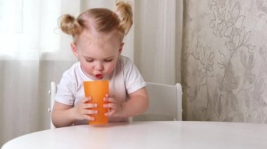 Beyaz tişörtlü küçük bir kız masada oturur ve bardaktan su içer.