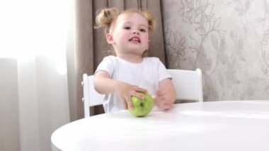 Beyaz tişörtlü küçük bir kız masada oturuyor ve yeşil elmayla oynuyor.