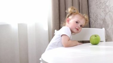 Beyaz tişörtlü küçük bir kız masada oturuyor ve yeşil elmayla oynuyor.