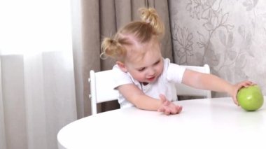Beyaz tişörtlü küçük bir kız masada oturuyor ve yeşil elmayla oynuyor.