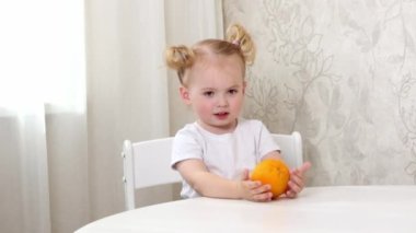 Beyaz tişörtlü küçük bir kız masaya oturur ve büyük bir portakalla oynar.