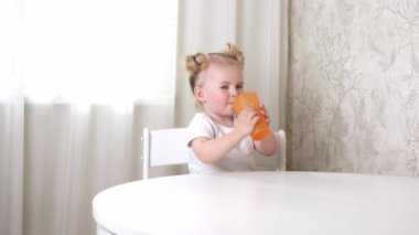 Beyaz tişörtlü küçük bir kız masada oturur ve bardaktan su içer.