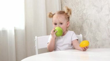 Beyaz tişörtlü küçük bir kız masada oturuyor ve meyvelerle oynuyor. limon, portakal ve elma