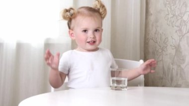 Beyaz tişörtlü küçük bir kız masada oturur ve bardaktan su içer.